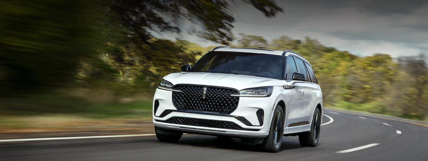 A white 2025 Lincoln Aviator® SUV is being driven down a road lined with trees. | Lincoln of Ramsey in Ramsey NJ