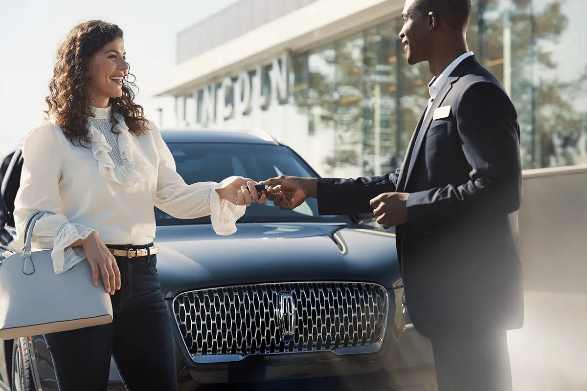 A dealership representative handing over car keys to a smiling customer in front of a Lincoln