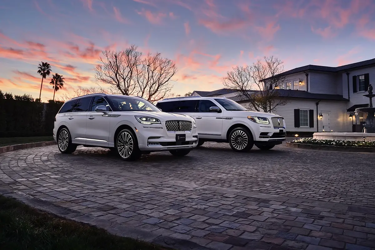 Two white Lincoln luxury SUVs parked on a cobblestone driveway in front of a large house at sunset.