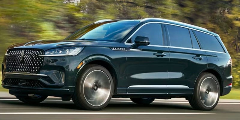 A dark blue Lincoln Aviator SUV drives on a road with green trees in the background.