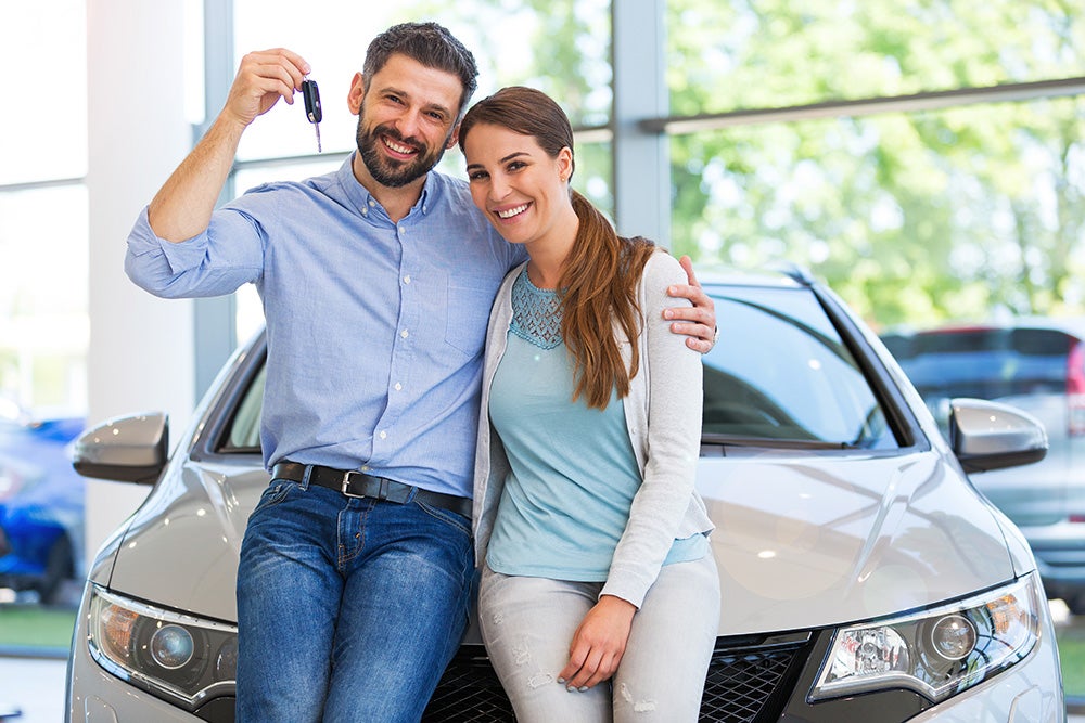 A smiling couple sitting on a car hood, holding up new keys