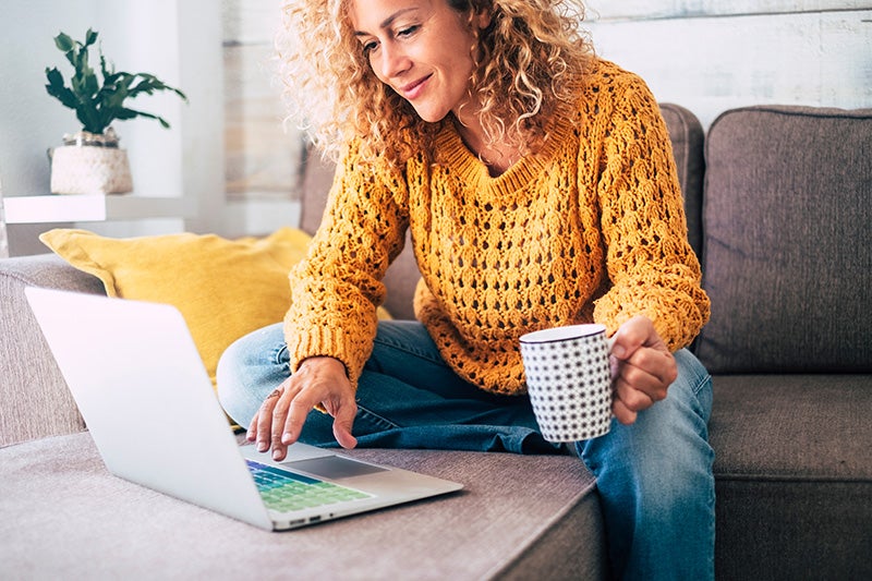 A curly-haired woman in a yellow sweater uses a laptop while holding coffee